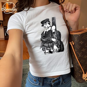 May include: White t-shirt featuring a black and white illustration of a character with a guitar. The character is wearing a hat, jacket, and skirt. The shirt is paired with blue jeans.