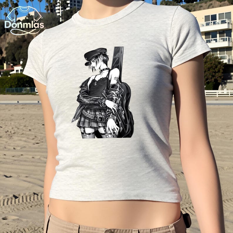 May include: A light gray t-shirt featuring a black and white illustration of a person with a guitar. The illustration depicts a person wearing a black hat, jacket, and plaid skirt. The shirt has a crew neck and short sleeves.