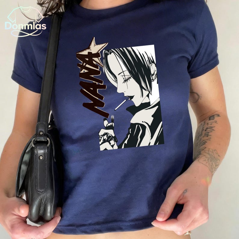 May include: Navy blue t-shirt with a black and white manga graphic of a person smoking, with the word "NANA" in a starburst design. The shirt has short sleeves.