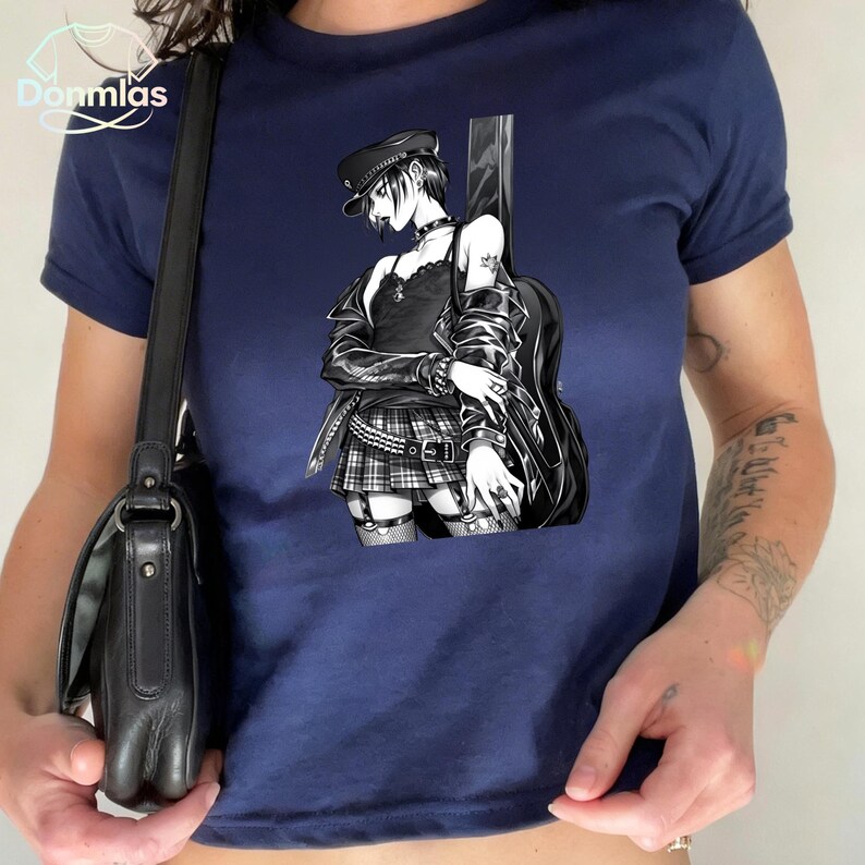 May include: Navy blue t-shirt featuring a black and white anime-style illustration of a person with a guitar. The person is wearing a black hat, jacket, and skirt. The shirt has the brand name "Donmlas" in the upper left corner.