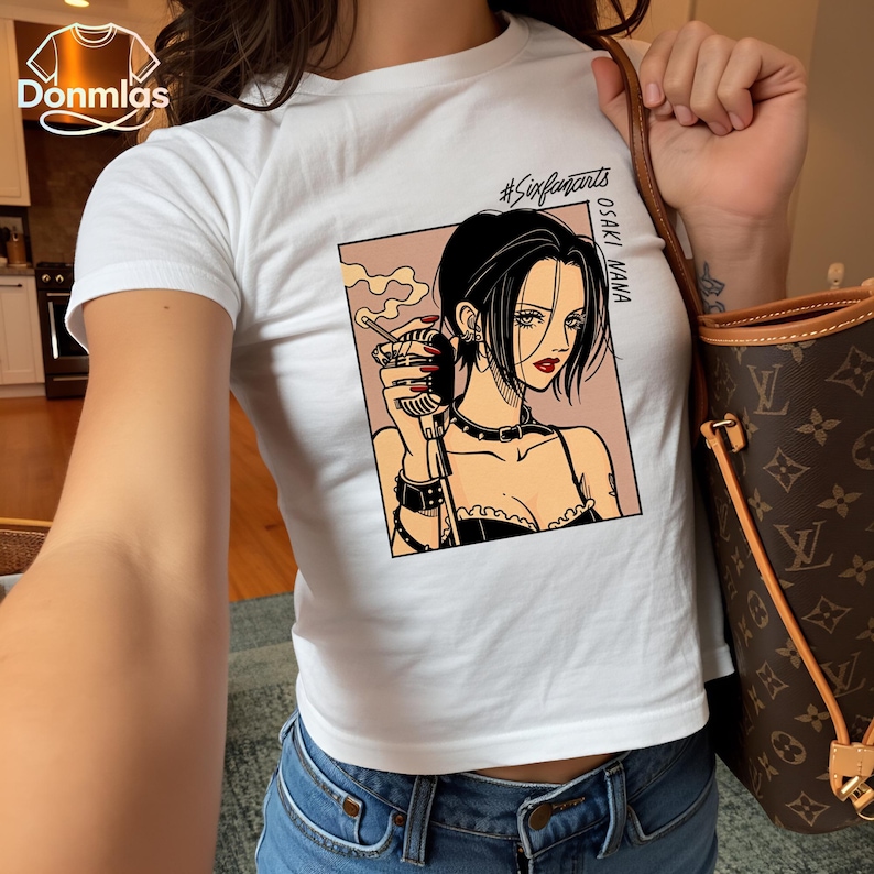 Puede incluir: Camiseta blanca con un gr&aacute;fico de una mujer de pelo negro, sosteniendo un micr&oacute;fono y un cigarrillo. El gr&aacute;fico incluye el texto "OSAKI NANA" y "#Sixfanarts". La camiseta se combina con unos vaqueros azules.