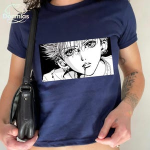 May include: Navy blue t-shirt featuring a black and white manga-style illustration of a person's face. The graphic is rectangular and centered on the shirt. The shirt has short sleeves and a crew neck.