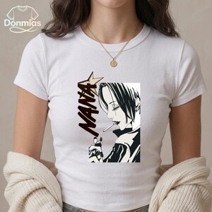 May include: White t-shirt with a black and white manga illustration of a person smoking. The word "NANA" is printed in a stylized font with a star. The shirt has short sleeves and a crew neck. The brand name "Donmlas" is visible.