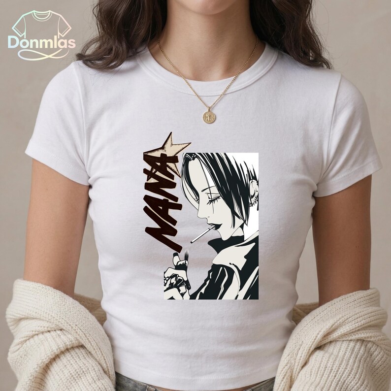May include: White t-shirt with a black and white manga illustration of a person smoking. The word "NANA" is printed in a stylized font with a star. The shirt has short sleeves and a crew neck. The brand name "Donmlas" is visible.