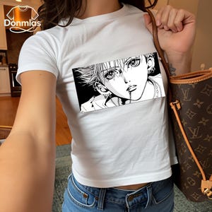 May include: White t-shirt featuring a black and white manga-style graphic of a person with multiple piercings and a chain in their mouth. The shirt is short-sleeved and worn with blue jeans.
