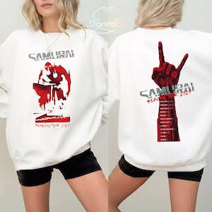 Könnte beinhalten: Weißes Sweatshirt mit roter und grauer Grafik. Auf der Vorderseite steht das Wort "SAMURAI" über einer roten Illustration und dem Text "REUNION TOUR 2023". Auf der Rückseite ist eine rote Handgeste und die Tourdetails abgebildet.