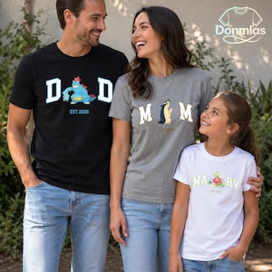 May include: A black t-shirt with a blue dinosaur graphic and the text DAD EST. 2026, a gray t-shirt with a yellow dinosaur graphic and the text MOM EST. 2026, and a white t-shirt with a green dinosaur graphic and the text MARY EST. 2026.