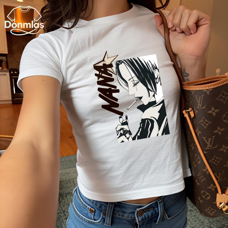 May include: White crop top with a manga-style graphic of a person smoking, with the word "NANA" in a bold font. The graphic is in black and white. The top is worn with blue jeans.