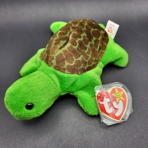 Könnte beinhalten: Ein grünes Plüschtier in Form einer Schildkröte mit einem braunen, gemusterten Panzer. Das Spielzeug hat ein schwarzes Auge und ein rotes und weißes Etikett. Auf dem Etikett steht "Beanie Baby" und der Markenname "Ty".