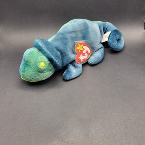 Può includere: Un camaleonte di peluche Beanie Baby verde e blu tie-dye con un'etichetta Ty rossa e bianca. L'etichetta ha una stella bianca e le parole "Ty Inc."