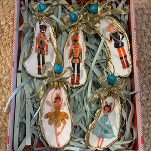 May include: Five oyster shell ornaments painted with nutcracker and ballerina figures. Each shell features a gold ribbon bow, a turquoise bead, and a gold hanging loop. The set is displayed in a white box filled with light blue paper shreds.