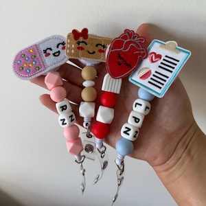 Puede incluir: Una colección de portatarjetas retráctiles con varios diseños. Los diseños incluyen una píldora sonriente, una tirita sonriente, un corazón rojo y una carpeta con una rodaja de sandía. Cada soporte tiene una cadena de cuentas con las letras RN o LPN.