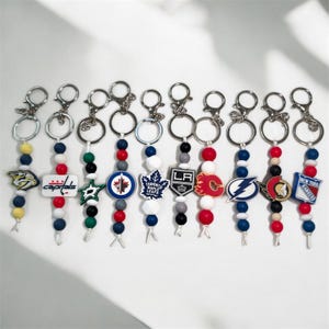 May include: A selection of keyrings displaying NHL team logos. Each keyring has a silver ring, a beaded strand in team colours, and a team logo charm. The keyrings are arranged on a white surface.