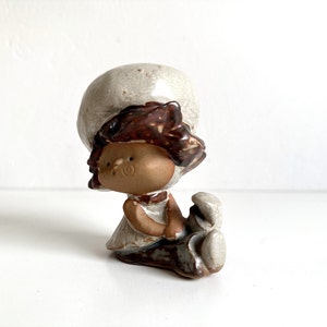 May include: A ceramic figurine of a girl with brown hair wearing a white hat and holding a small ceramic animal. The figurine is painted in shades of brown, white, and beige.
