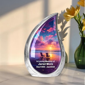May include: A silver teardrop-shaped memorial urn featuring a sunset beach scene. The urn has engraved details and the text "In Loving Memory of Jared More Dec/1956 - Jan/2025."