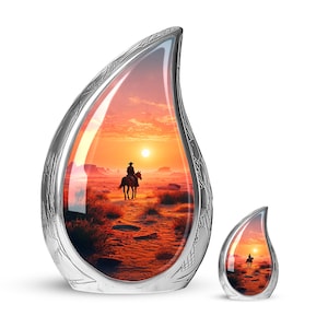 May include: Two silver teardrop-shaped urns. The larger urn features a sunset scene with a cowboy on horseback. The smaller urn mirrors the same image. Both urns have a detailed, engraved silver exterior.
