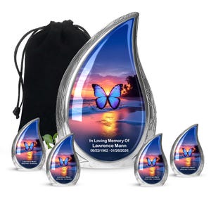 May include: A set of memorial urns with a butterfly theme. The main urn is teardrop-shaped with a silver frame and a sunset scene with a butterfly. Smaller matching urns are included, along with a black drawstring bag. Text reads "In Loving Memory Of Lawrence Mann 09/22/1962 - 01/26/2026."