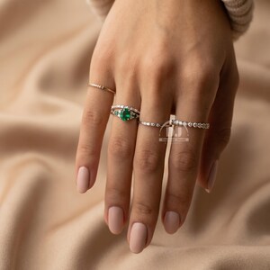May include: A hand adorned with multiple rings. One ring showcases a large, oval emerald gemstone, encircled by smaller diamonds. Other rings include slender bands set with tiny, sparkling stones. The rings are displayed against a neutral backdrop.