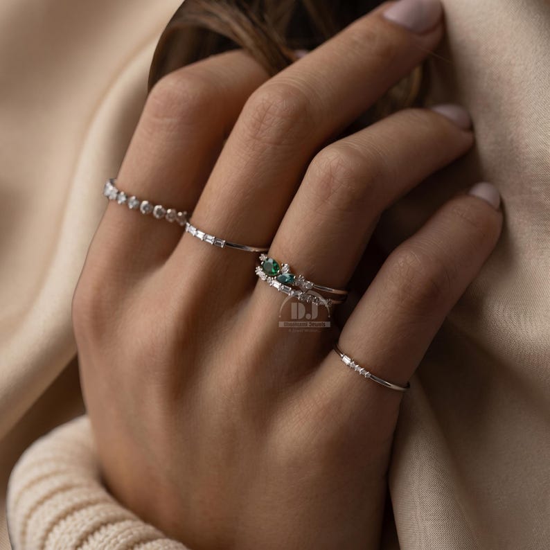 May include: A hand shows off multiple silver rings. One ring has a row of small, clear stones. Another has a line of rectangular stones. A third ring features a cluster of green stones. The fourth ring has a few small, clear stones.
