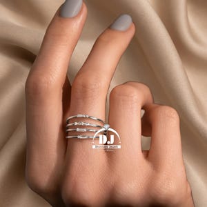 May include: A silver ring featuring four bands, each set with small, clear gemstones. A larger, diamond-shaped gem is at the top. The ring is on a finger with grey nail polish. The logo "Dhanlaxmi Jewels" is visible.