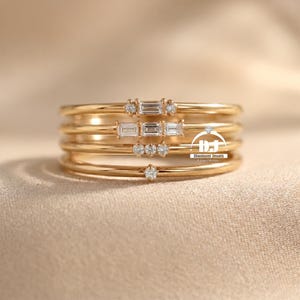 May include: Five gold rings are stacked, each with a slender band. Some rings are adorned with small, clear gemstones, either round or rectangular. The rings are presented on a neutral-toned surface, creating a simple and elegant display.