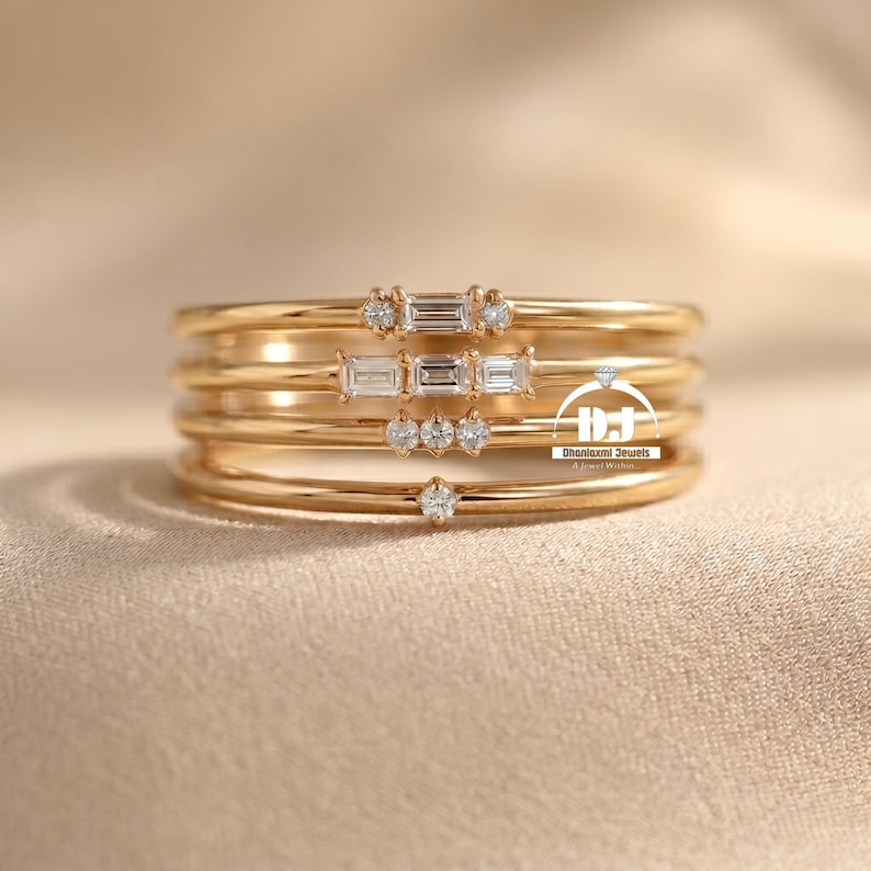 May include: Five gold rings are stacked, each with a slender band. Some rings are adorned with small, clear gemstones, either round or rectangular. The rings are presented on a neutral-toned surface, creating a simple and elegant display.