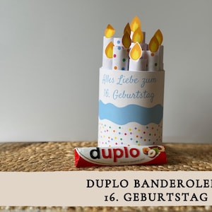 May include: A cylindrical birthday decoration with paper candles and flames. The cylinder is white with a blue wave design and colorful polka dots. The text "Alles Liebe zum 16. Geburtstag" is visible. A red and white candy bar is in front.