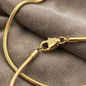 Op de afbeelding: Een goudkleurige slangenketting met een gepolijste karabijnsluiting. De ketting heeft een gladde, flexibele en afgeronde textuur met een doorlopend in elkaar grijpend schakelpatroon dat zorgt voor een strakke en uniforme uitstraling.