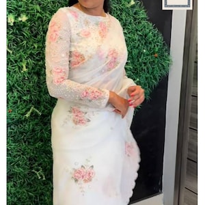 May include: A white saree with floral embroidery. The saree has a sheer overlay with delicate pink rose designs on the sleeves and body. The text "Siyaposhak" is visible at the bottom of the image.