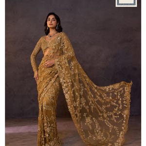 May include: A golden-yellow sari with detailed floral embroidery. The sari has a sheer, flowing fabric with long sleeves and a deep V-neckline. The brand name "Siyaposhak" is visible at the bottom of the image.