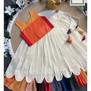 Op de afbeelding: Een crèmekleurige rok en een bijpassende oranje en rode top. De rok heeft een geschulpte zoom met een regenboog aan kleuren. De top is mouwloos. De afbeelding bevat de tekst "Siyaposhak".