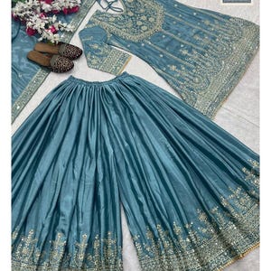 Może przedstawiać: Trzyczęściowy, haftowany strój w kolorze morskim. Zestaw zawiera tunikę z długim rękawem, spodnie z szerokimi nogawkami i pasujący szal. Tunika i spodnie mają skomplikowane złote hafty. Widoczna jest nazwa marki "Siyaposhak".