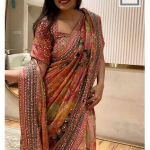 Peut inclure: Une femme porte un sari coloré avec des motifs complexes dans des tons de rouge, orange et vert. Le sari présente des broderies et des ornements détaillés. Le texte "Siyaposhak" est visible sur le vêtement.