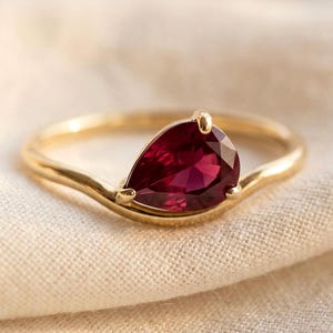 Könnte beinhalten: Ein Goldring mit einem einzelnen birnenförmigen, tiefroten Edelstein in einer Krappenfassung. Die Ringschiene hat eine glatte, polierte Oberfläche und eine sanfte Kurve, die der Form des tropfenförmigen Schliffs folgt und ein schlichtes Design bildet.