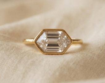 May include: A gold ring with a large, clear, emerald-cut gemstone. The gemstone is set in a hexagonal bezel. The ring band is thin and gold. The ring is displayed on a cream-colored fabric.