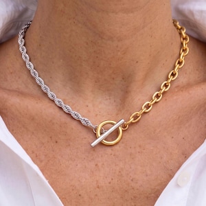 May include: A two-tone necklace featuring a silver-coloured rope chain on one side and a gold-coloured link chain on the other. The necklace is secured at the front with a gold-coloured circular toggle ring and a silver-coloured bar closure.