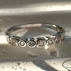May include: A silver ring featuring alternating square and round clear gemstones. The ring is displayed on a white surface and has the text "Siena ET MOI" in cursive script.