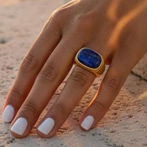 May include: A gold ring with a large, rectangular, deep blue stone. The ring is on a hand with white nail polish. The ring is a statement piece, perfect for adding a pop of color to any outfit.