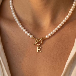 May include: A pearl necklace with a gold clasp and a gold letter "E" pendant. The necklace is worn against a white button-down shirt. The pearls are small and round, and the clasp is a simple bar and loop design.