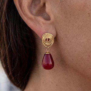 May include: A gold-toned earring featuring a circular, wire-wrapped stud component connected to a smooth, teardrop-shaped deep red stone pendant. The earring is suspended from the earlobe, showcasing a polished metallic finish and a vibrant gemstone.