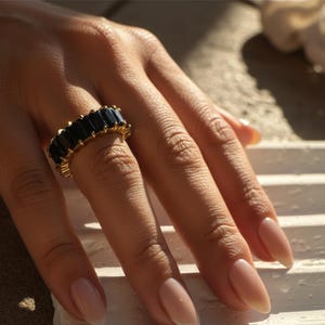 May include: A gold-toned ring with a row of rectangular, dark blue gemstones. The ring is on a hand with long, polished nails. The ring's design is simple and elegant, suitable for everyday wear or special occasions.