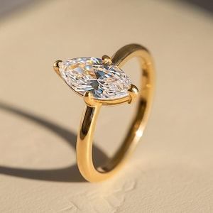May include: A gold ring with a marquise-cut diamond. The diamond is held in place by four prongs and reflects light. The band is made of polished gold. The ring is shown against a plain background, emphasizing its sophisticated design.