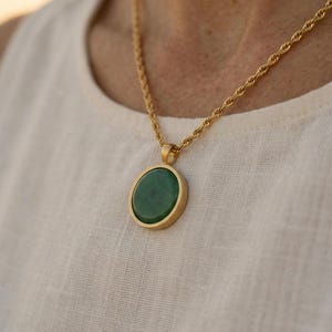 Op de afbeelding: Een goudkleurige ketting met een cirkelvormige groene hanger. De hanger is gezet in een gouden frame en hangt aan een bijpassende gouden ketting. De ketting heeft een gedraaid touwontwerp. De ketting is een sieraad.