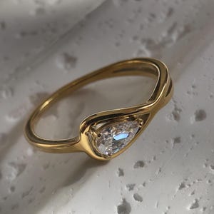 May include: A gold ring featuring a pear-cut diamond. The ring's band has a distinctive, flowing design that holds the diamond. The diamond sparkles, reflecting light. The ring is set against a neutral background.