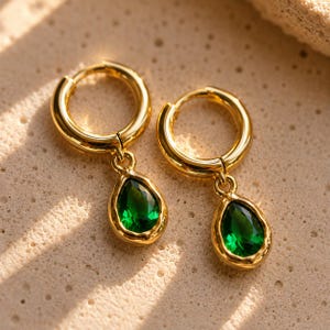 May include: A pair of gold hoop earrings featuring dangling teardrop-shaped pendants. Each pendant contains a faceted, deep green gemstone set within a polished gold frame. The hoops are circular and smooth, connecting to the teardrop charms.