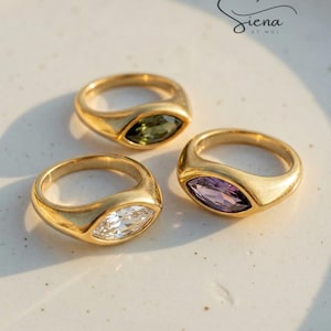 Può includere: Tre anelli dorati con pietre preziose taglio marquise, verdi, bianche e viola. Gli anelli hanno una finitura lucida e sono su una superficie bianca. Fanno parte della collezione "Siena by Moi".