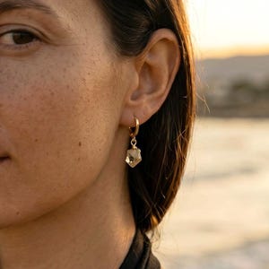 May include: A gold hoop earring featuring a faceted, pale gemstone pendant. The earring is displayed on a person's ear, with the jewellery as the focal point. The background is blurred, implying an outdoor location.