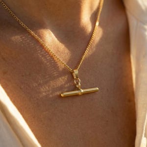Peut inclure: Un collier doré avec un pendentif en forme de barre en T. La chaîne est fine et le pendentif est une barre simple avec une petite sphère centrale. Le collier est porté contre la peau.