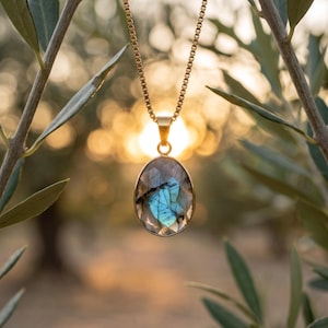 May include: A gold-toned necklace featuring an oval-shaped labradorite pendant with a faceted surface. The stone displays a prominent iridescent blue flash against a grey base. The pendant hangs from a delicate gold-toned box chain link necklace.