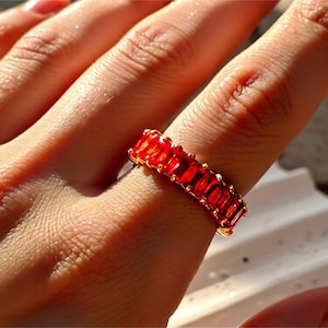 May include: A gold ring featuring a band of vivid red rectangular gemstones. The ring is displayed on a finger, highlighting the detailed design and colour. The gemstones are closely set, forming a continuous, striking pattern. The ring's design is elegant.